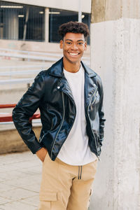 Young cheerful black male in leather jacket standing on street with hands in pockets near column and looking at camera