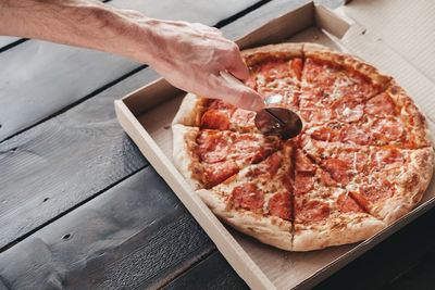 Male hand cuts pepperoni pizza with a round knife on a dark wooden background. 