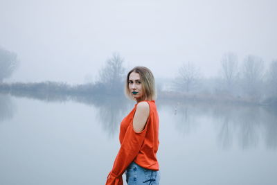Portrait of young woman standing by lake against sky during winter