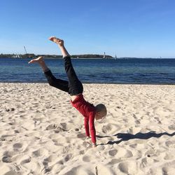 Full length of girl performing handstand at beach against clear blue sky