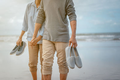 Midsection of couple holding footwears while walking on shore at beach