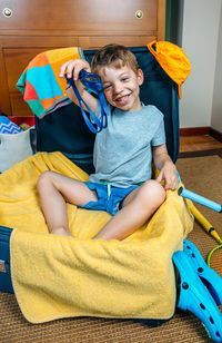 Portrait of smiling boy holding swimming goggles while sitting in suitcase at home