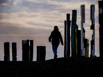 Silhouette woman walking by wooden posts on field against cloudy sky