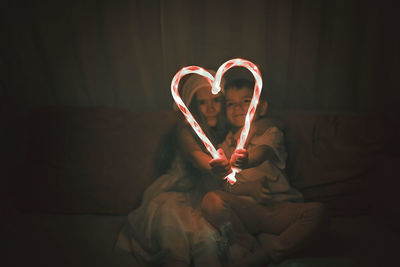 Siblings making heart shape of illuminated candy canes on sofa in darkroom