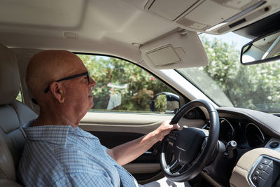 Side view of concentrated senior man in checkered shirt and eyeglasses driving modern car on sunny day in countryside