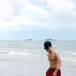 Shirtless young man walking on shore at beach against cloudy sky