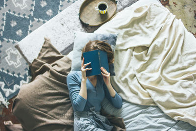 High angle view of woman holding book against face while lying on bed at home