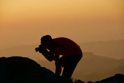 Silhouette man photographing while standing on mountain against sky during sunset