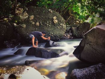 Side view of man photographing while standing in stream against rocks