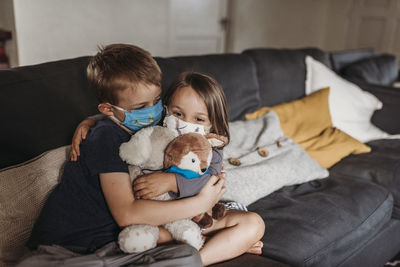 Young girl and school-age boy with masks hugging and smiling on couch