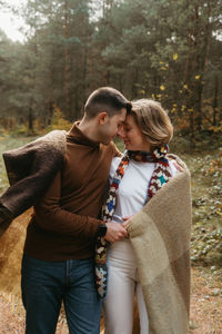 A man and a woman covered in a blanket snuggled up to each other in the middle of the forest