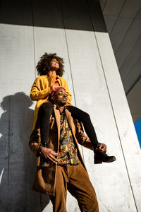 From below of modern african american guy in sunglasses and jacket carrying trendy black girlfriend on shoulders standing on street looking away