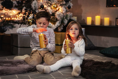 Children holding candles and making wish, sitting on floor in dark room. christmas tree  background