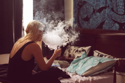 Blond woman smoking while sitting in bedroom