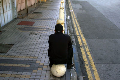 Rear view of man sitting on bollard at footpath