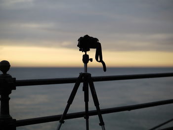 Close-up of silhouette camera on railing against sky during sunset