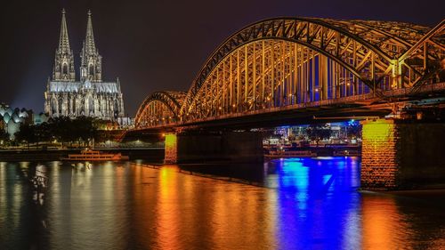 Illuminated hohenzollern bridge over rhine river by cologne cathedral against sky at night