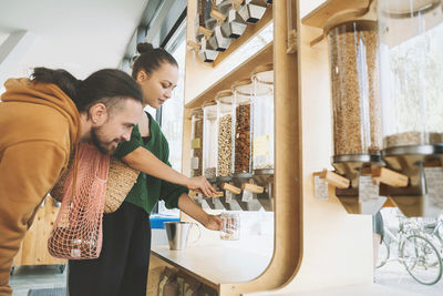 Man looking at woman filling mason jar under food dispenser at zero waste store