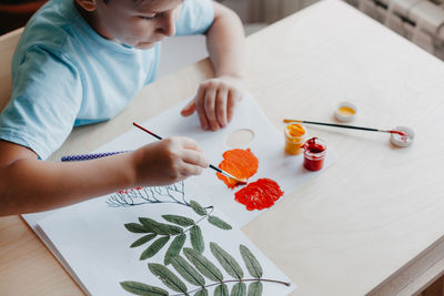 Cute child boy siting at desk and drawing rowanberries on album sheet with dry rowan leaves. autumn