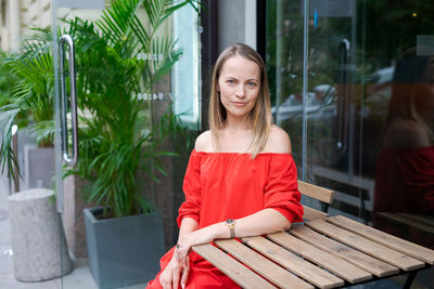 A confident young woman sits at a table in a street cafe in a red dress