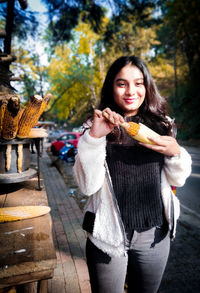 Portrait of smiling young woman having corn at market stall in city