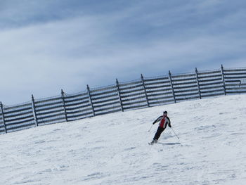 Person skiing on snow covered field by fence against cloudy sky