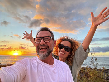 Portrait of smiling friends with arms raised standing against sky during sunset