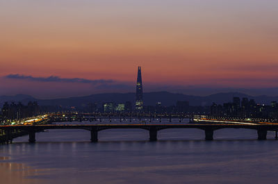 Illuminated bridge over river by buildings against sky at sunset