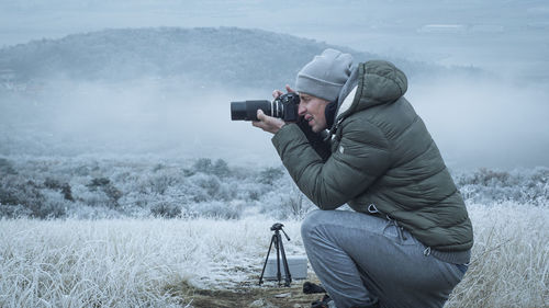 Side view of man photographing while crouching on field during winter