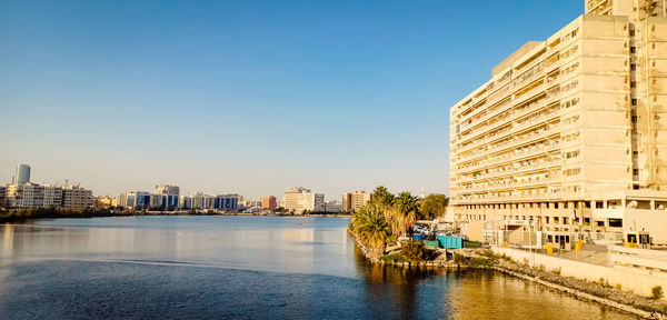Modern buildings by river against clear blue sky