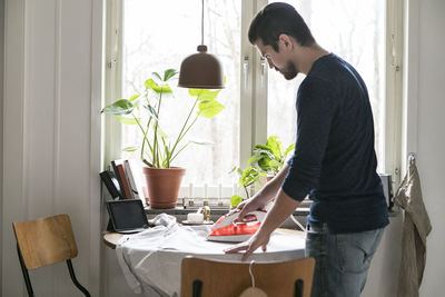 Side view of young man standing by window at home