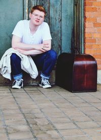 Full length portrait of young man with suitcase sitting outside closed door