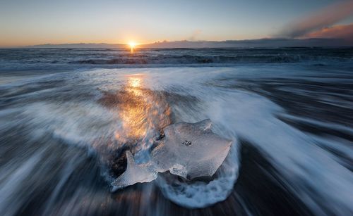 Blurred motion of wave with ice on shore against sky during sunset
