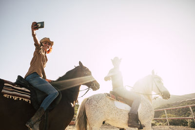 Low angle view of young woman taking selfie with man while riding horses against sky during sunny day