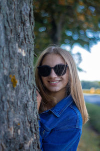 Portrait of young woman wearing sunglasses standing by tree trunk