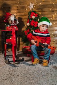 Smiling boy sitting with toy guitar in santa hat with wooden wall background and christmas tree