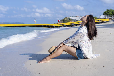 Worried woman sitting on beach with a straw hat on her knees beach by the sea in sunny day time.