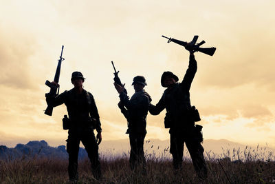 Silhouette army soldiers holding guns while standing on field against sky during sunset