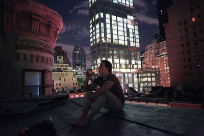 Young man drinking beer on house roof against illuminated modern building