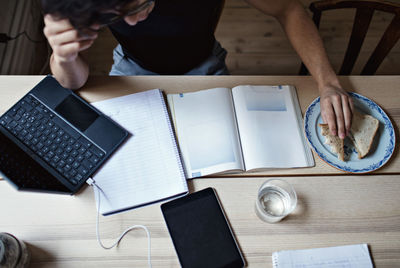 High angle view of dedicated young man doing homework while having sandwich at table