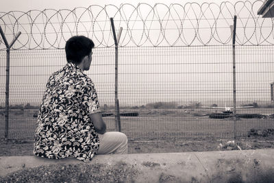 Rear view of man sitting against chainlink fence