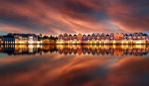 Illuminated buildings by lake against sky during sunset
