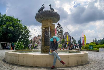 Portrait of woman wearing hat sitting on retaining wall against fountain