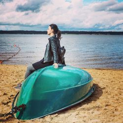 Woman sitting on upside down boat on shore at beach against cloudy sky