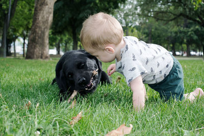 Cute toddler boy playing with black dog on relaxing on grassy field