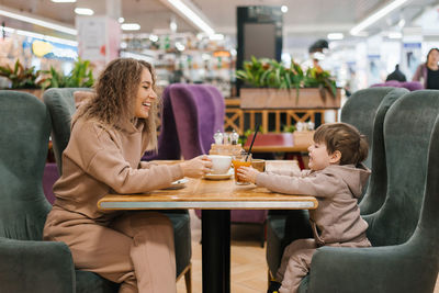 Young curly haired mother and her little son are having breakfast or lunch in a cafe