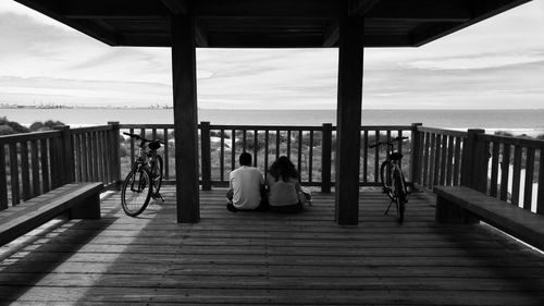 People sitting on railing by sea against sky