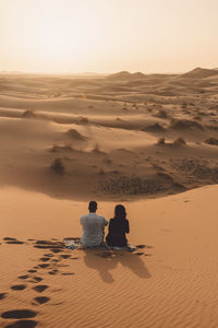 Rear view of friends sitting on sand at sahara desert against sky during sunset