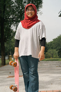 Young girl wearing hijab is running while holding pennyboard in the park. view from behind.