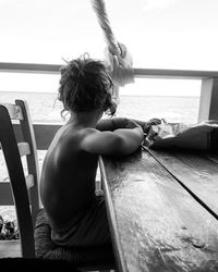 Side view of shirtless boy sitting at table on boat in sea